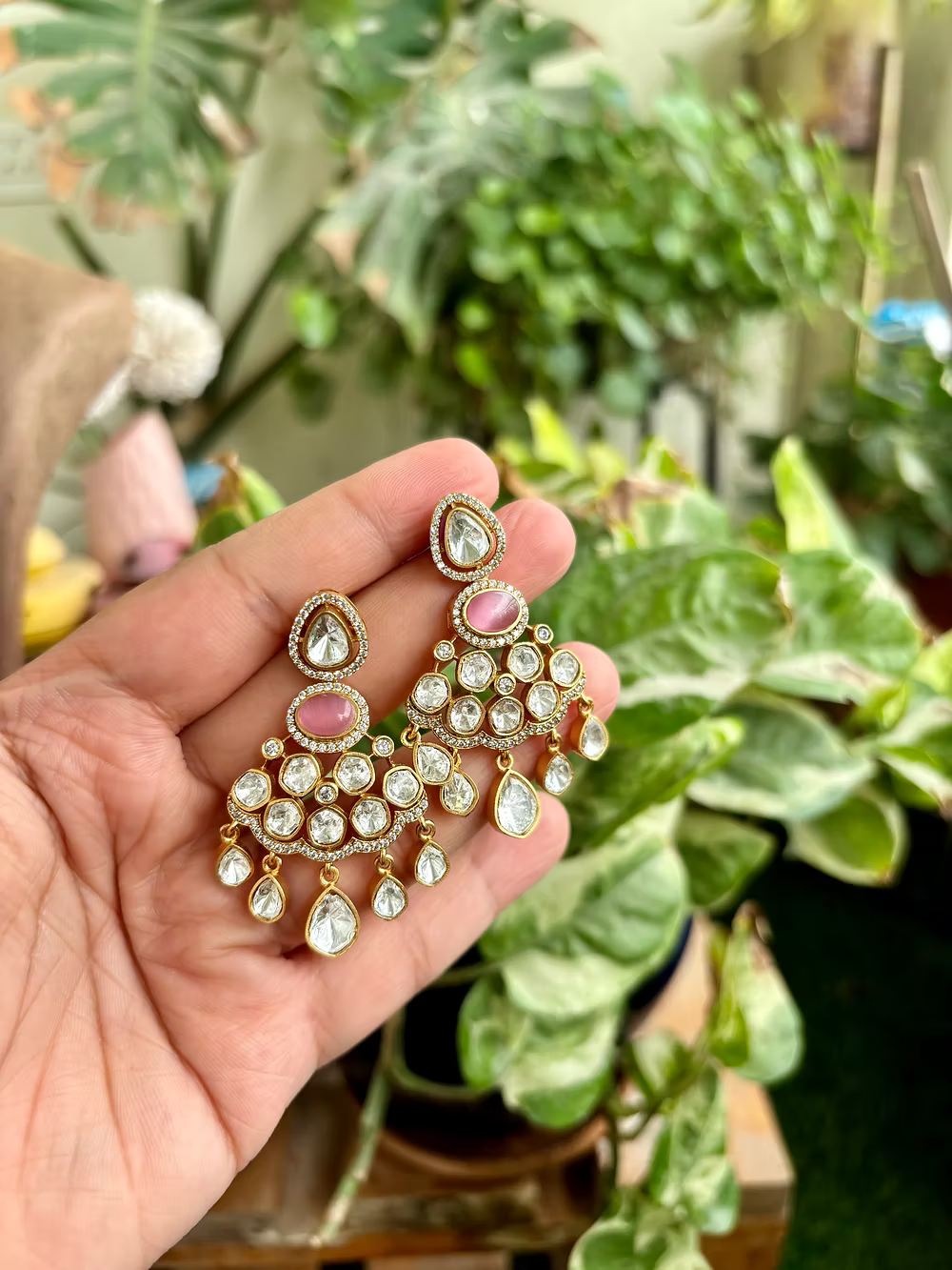 Gold and pink earrings held in a hand with a blurred plant background