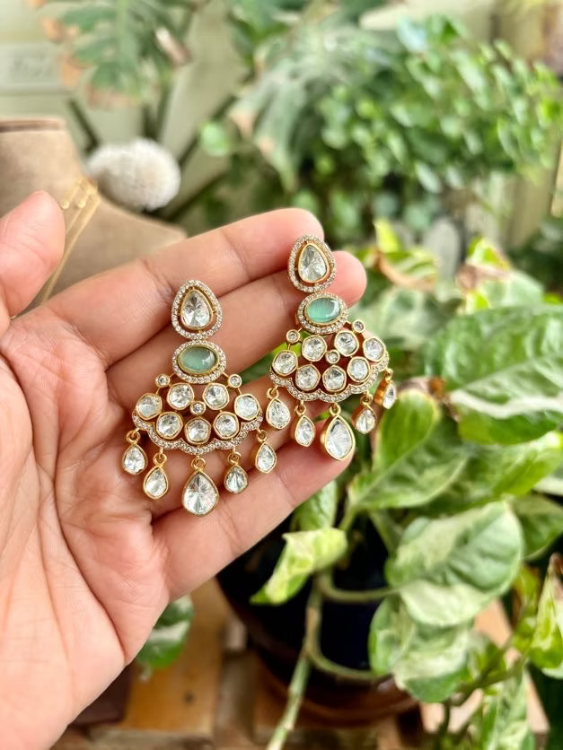 Delicate gold and gemstone earrings held in a hand with a green plant background